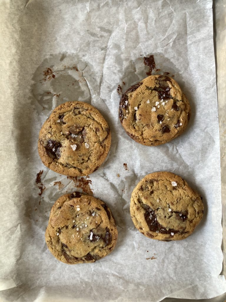 Four chocolate chip cookies on a baking tray, with sea sat on top and chocolate smears